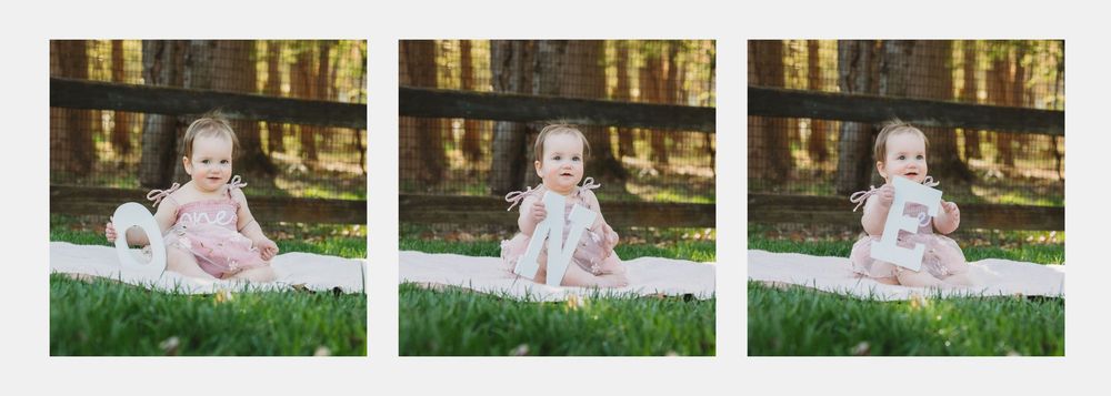 A baby girl in a pink dress sits on a white blanket in green grass near a wooden fence in a sunny outdoor setting.