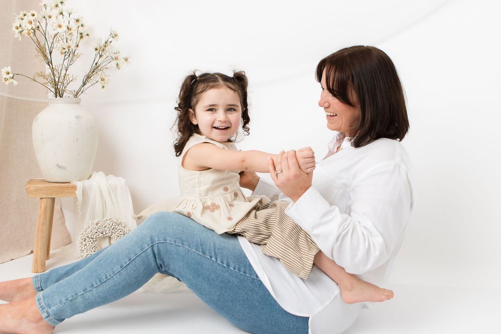 A parent and child in casual attire play together while sitting on a white floor with a decorative vase in background.