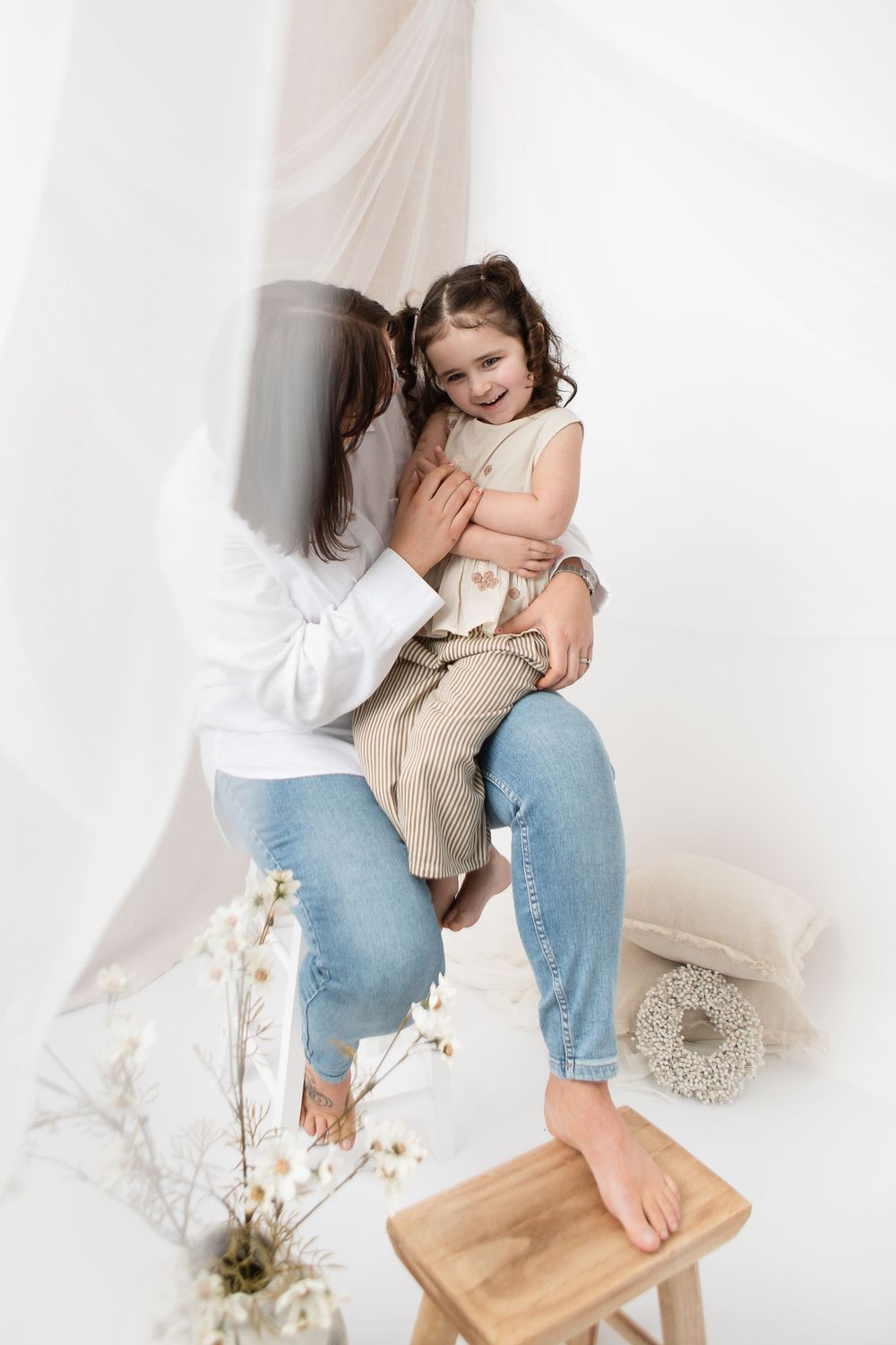 Intimate lifestyle portrait featuring tender family moment against white curtain backdrop with wooden stool and floral decor.