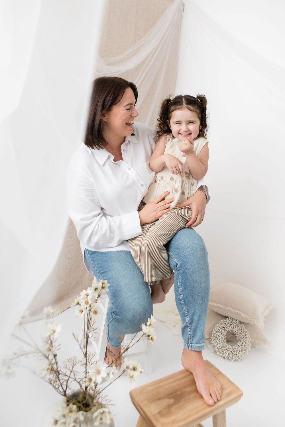 Two people share joyful laughter together while sitting against a white backdrop with dried flowers nearby.
