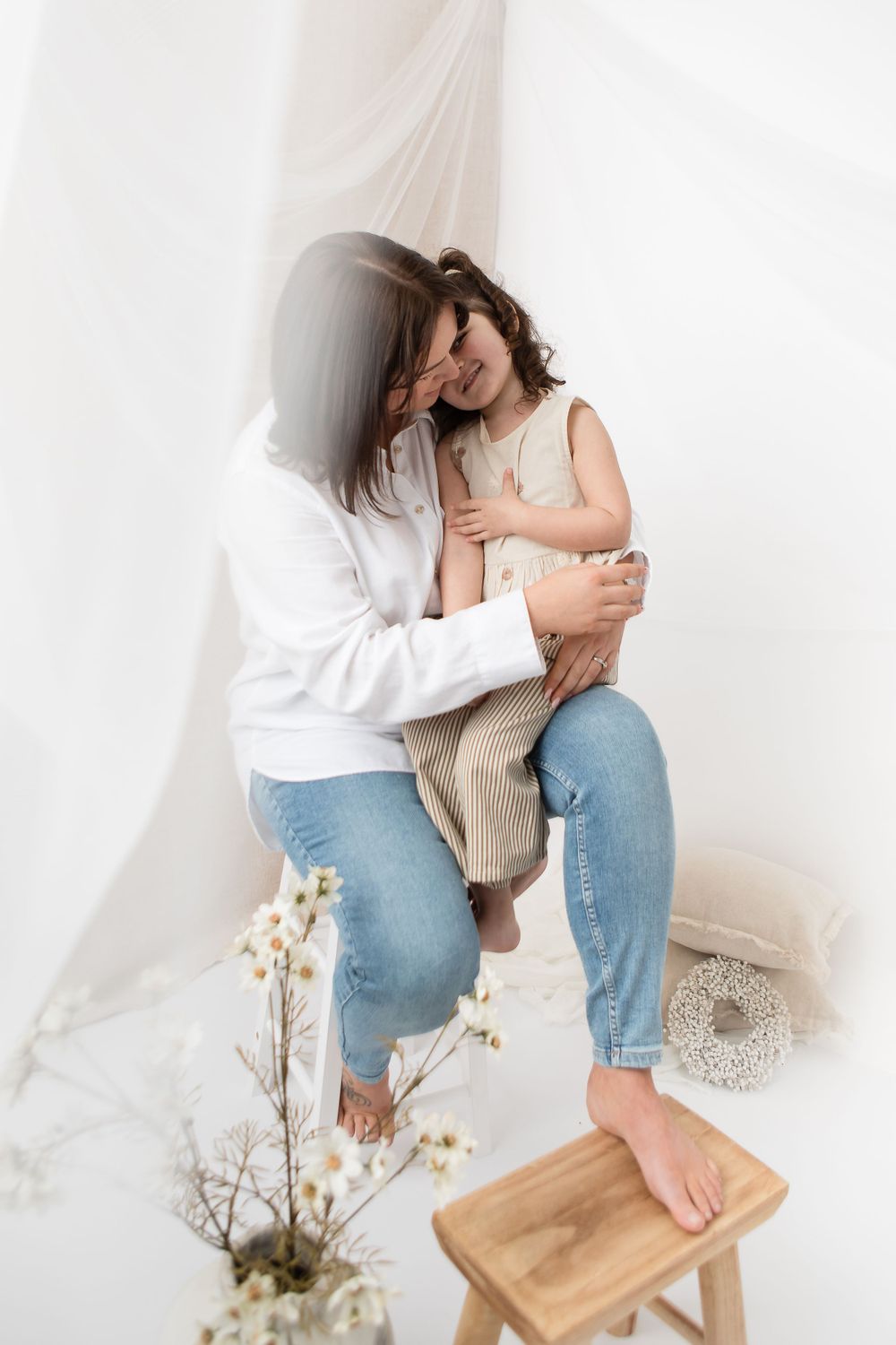 Mother and child sharing sweet moment together against white curtain with wooden stool and dried flowers nearby.