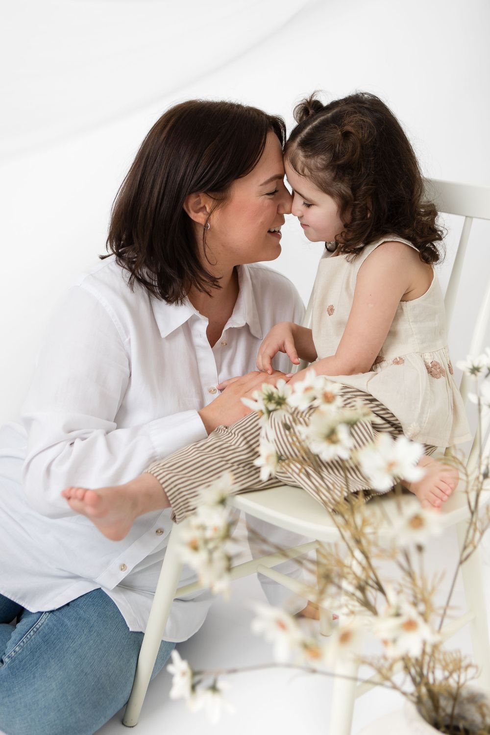 Two people share a tender moment while sitting by white flowers in a bright minimalist setting.