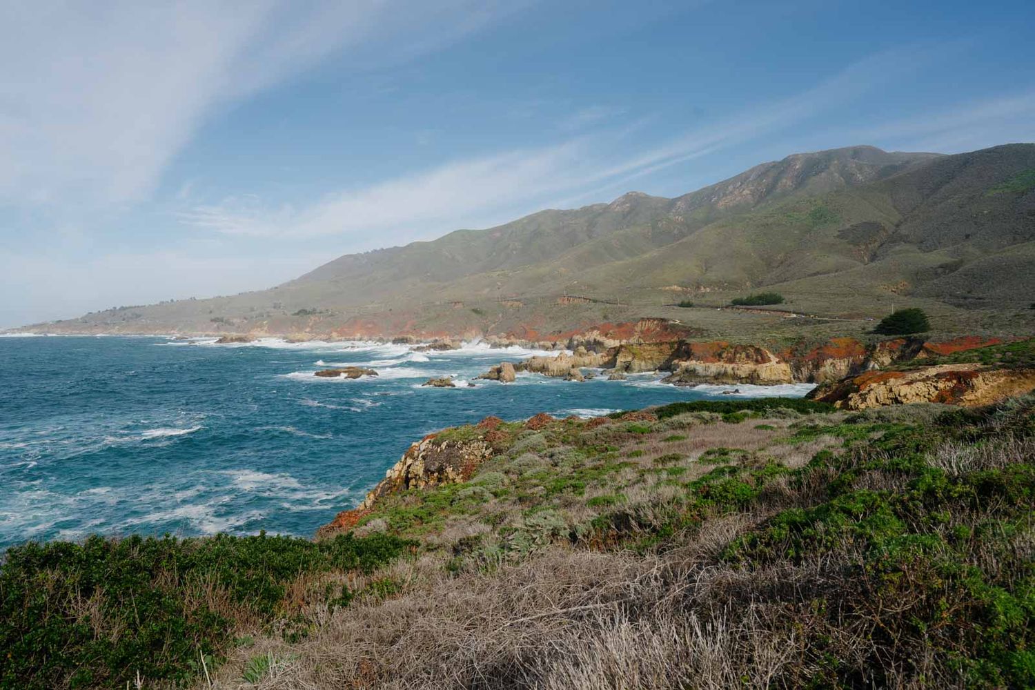 Rugged coastline of Big Sur California with waves crashing against rocky cliffs under blue skies.
