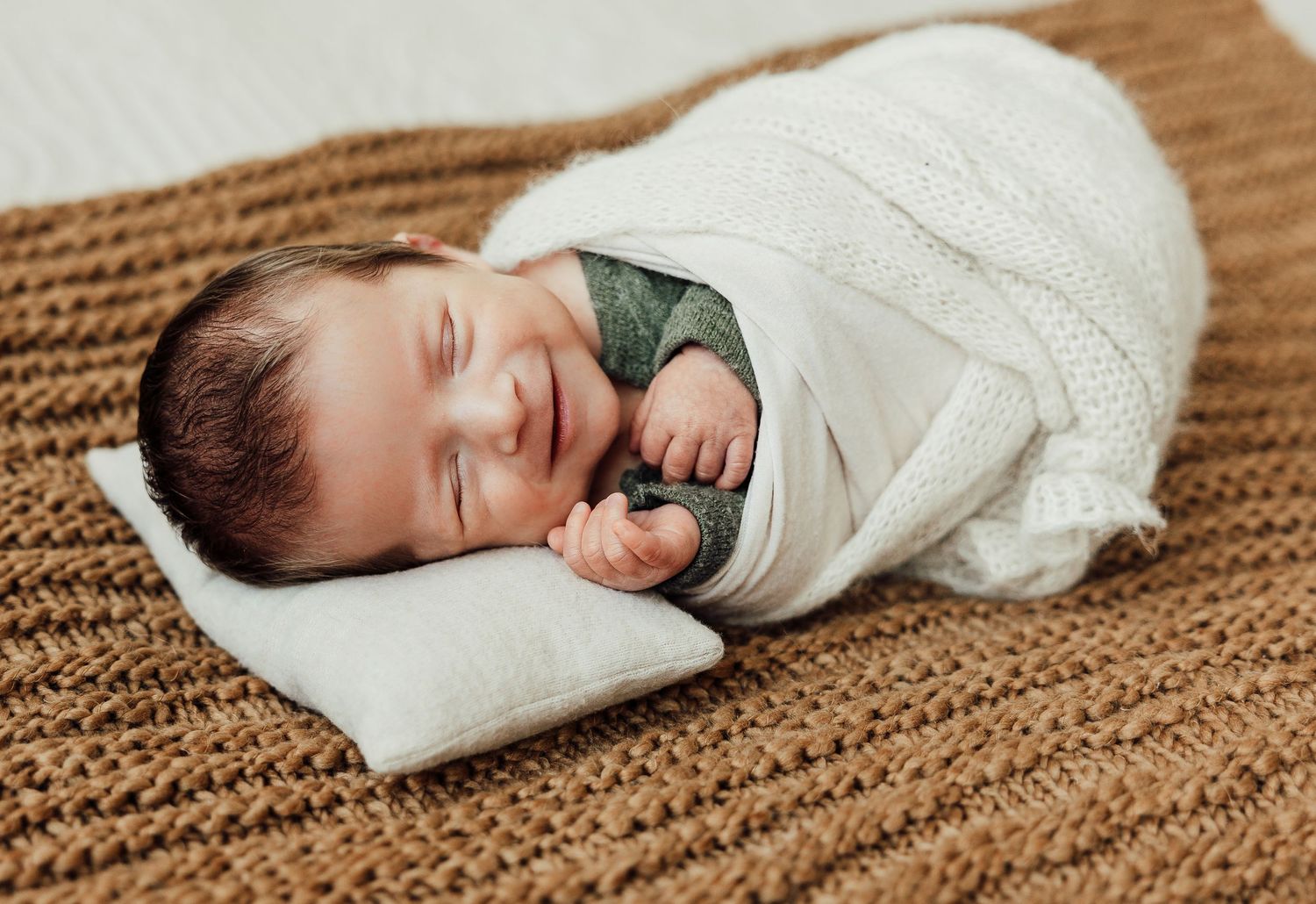 Sleeping newborn wrapped in white swaddle on textured brown knit blanket.