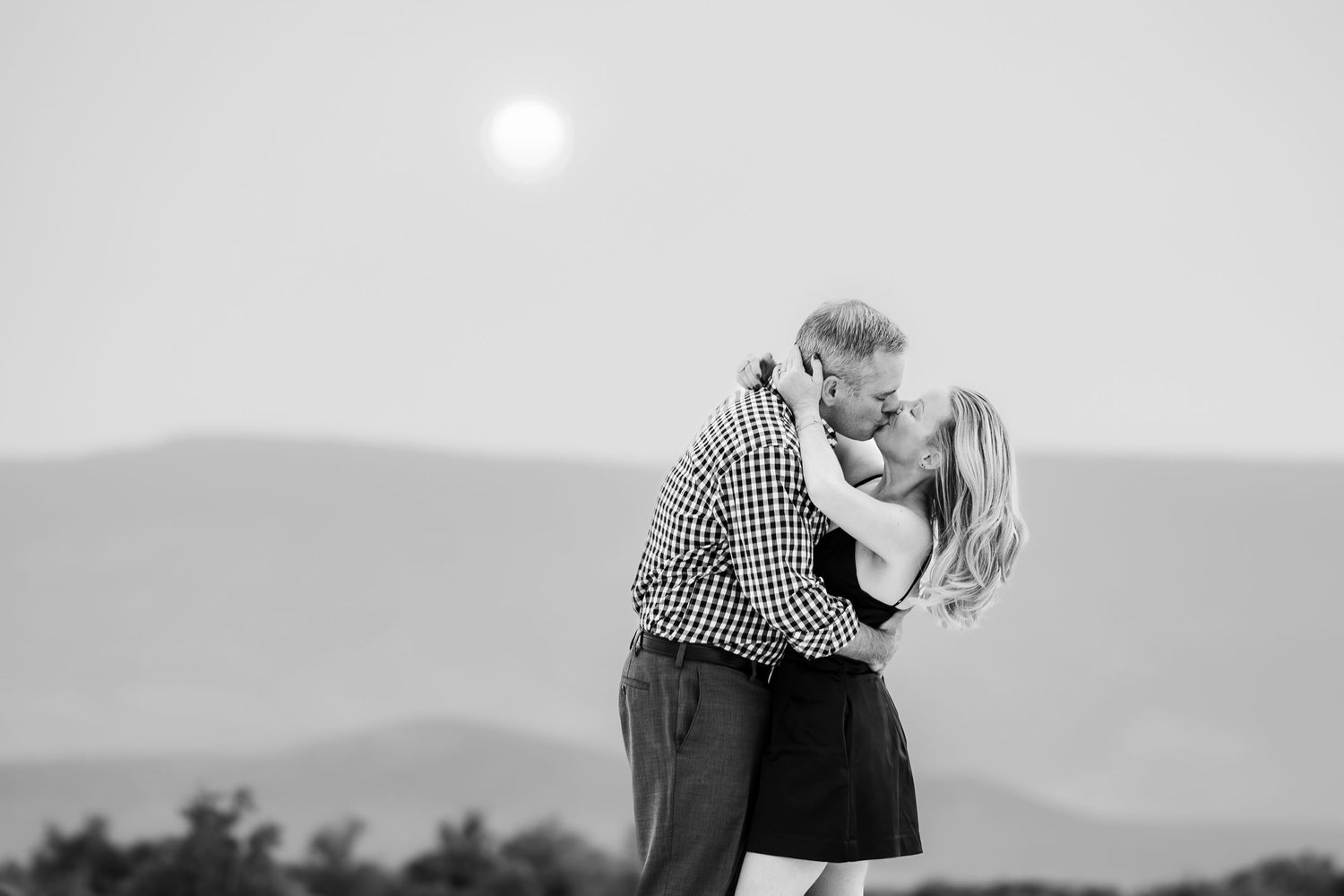 An intimate black and white portrait of a couple sharing a romantic moment against mountain scenery.