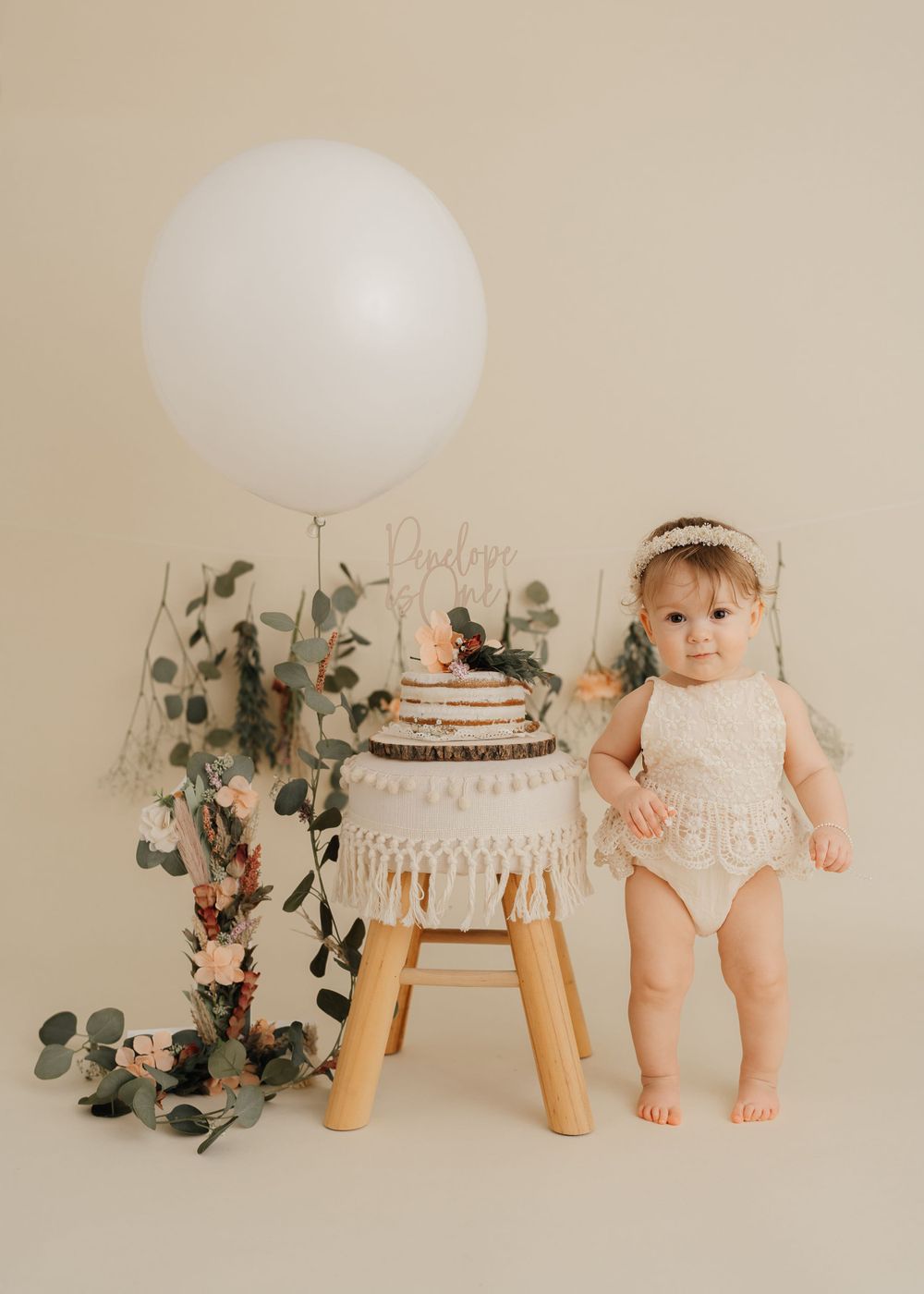 Baby in lace outfit standing beside wooden stool with cake and white balloon against floral backdrop.