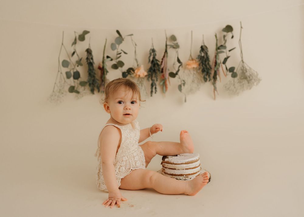 Baby in lace romper sitting by rustic cake with eucalyptus garland backdrop.