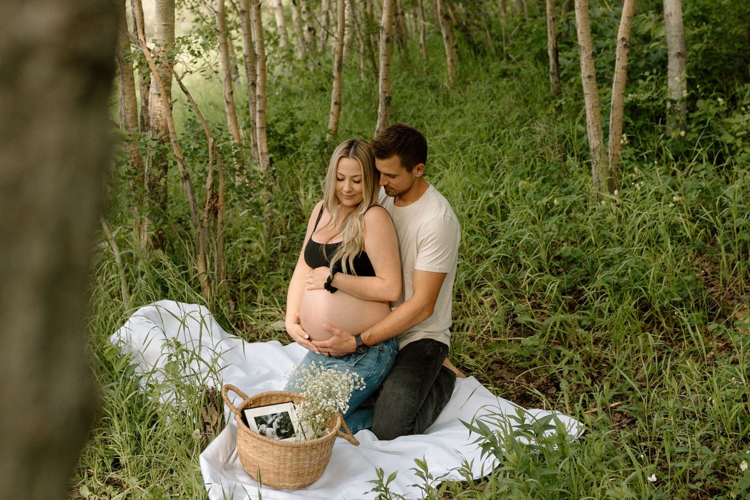 A romantic maternity picnic scene on a blanket in a wooded forest setting with a wicker basket.