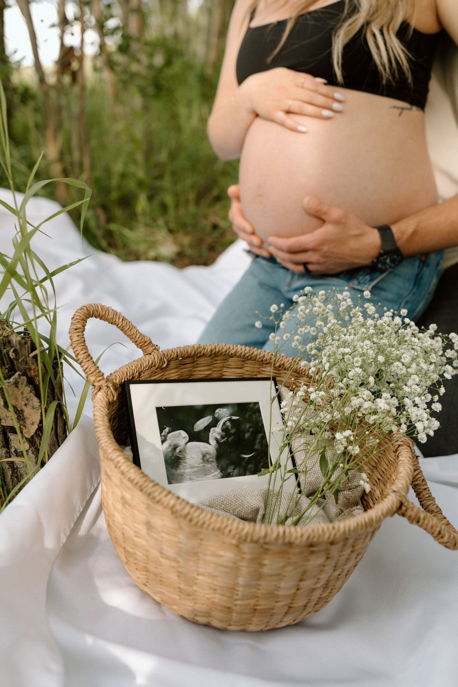 Pregnancy photoshoot detail with basket containing ultrasound photo and baby's breath flowers on white blanket.