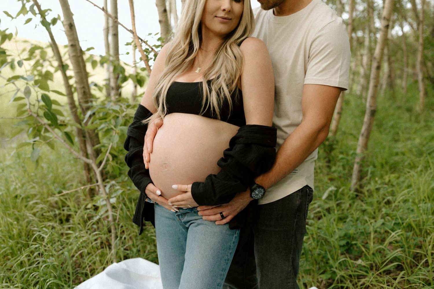 Couple embraces in a sun-dappled forest setting during pregnancy photoshoot with her wearing black crop top and jeans.