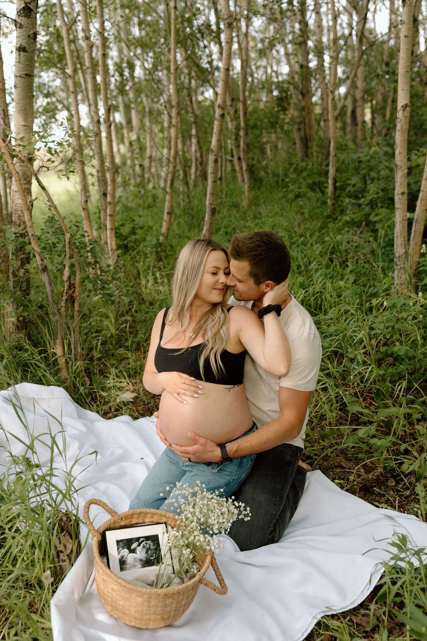 A romantic maternity photo shoot in a wooded area with a white blanket and floral basket containing an ultrasound photo.