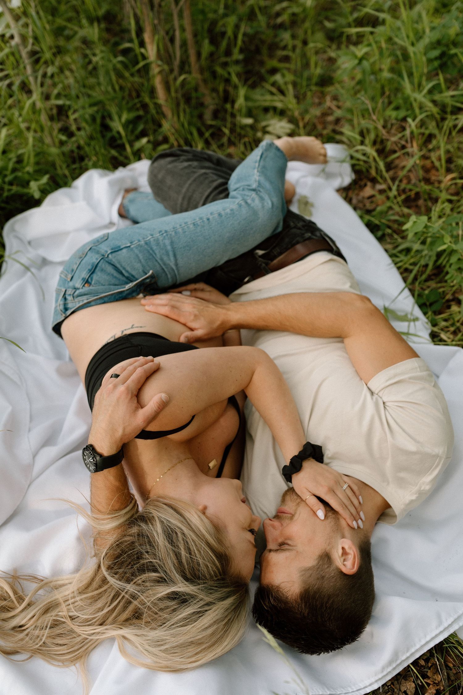Couple lying on white blanket outdoors sharing intimate moment among tall grass and greenery.