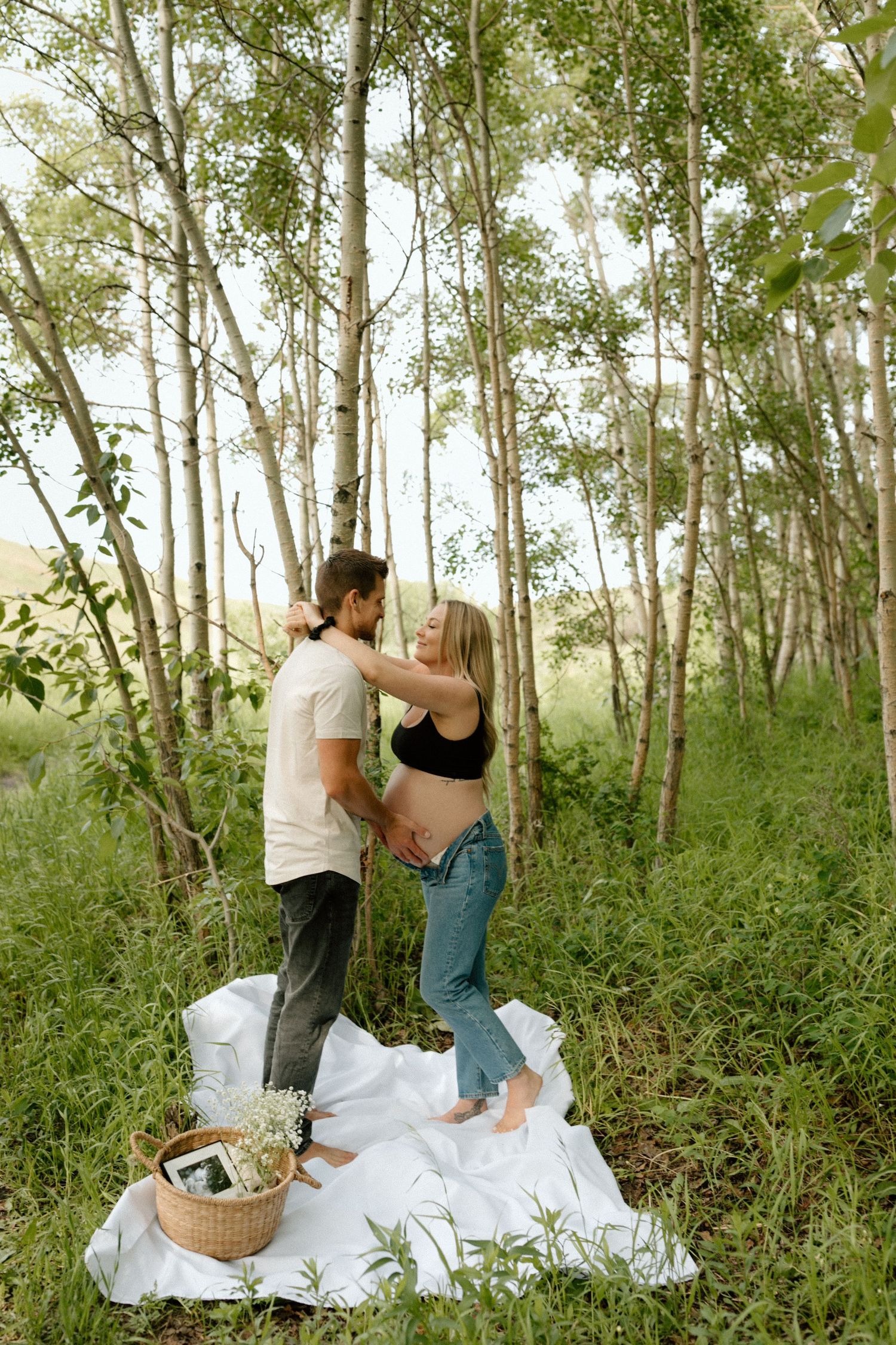 A maternity photography session in a sunlit birch grove with a white picnic blanket setup.