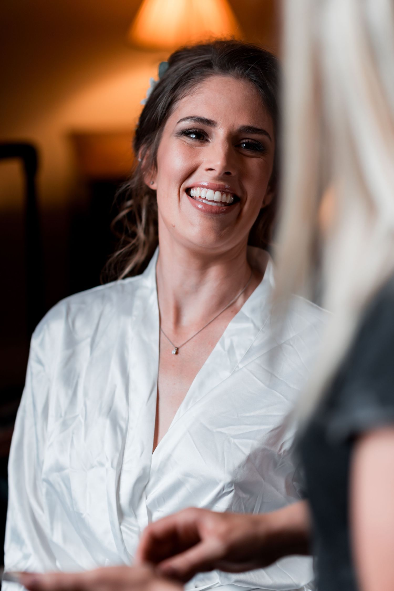 Woman wearing white silk robe smiles warmly while getting ready in warm indoor lighting.