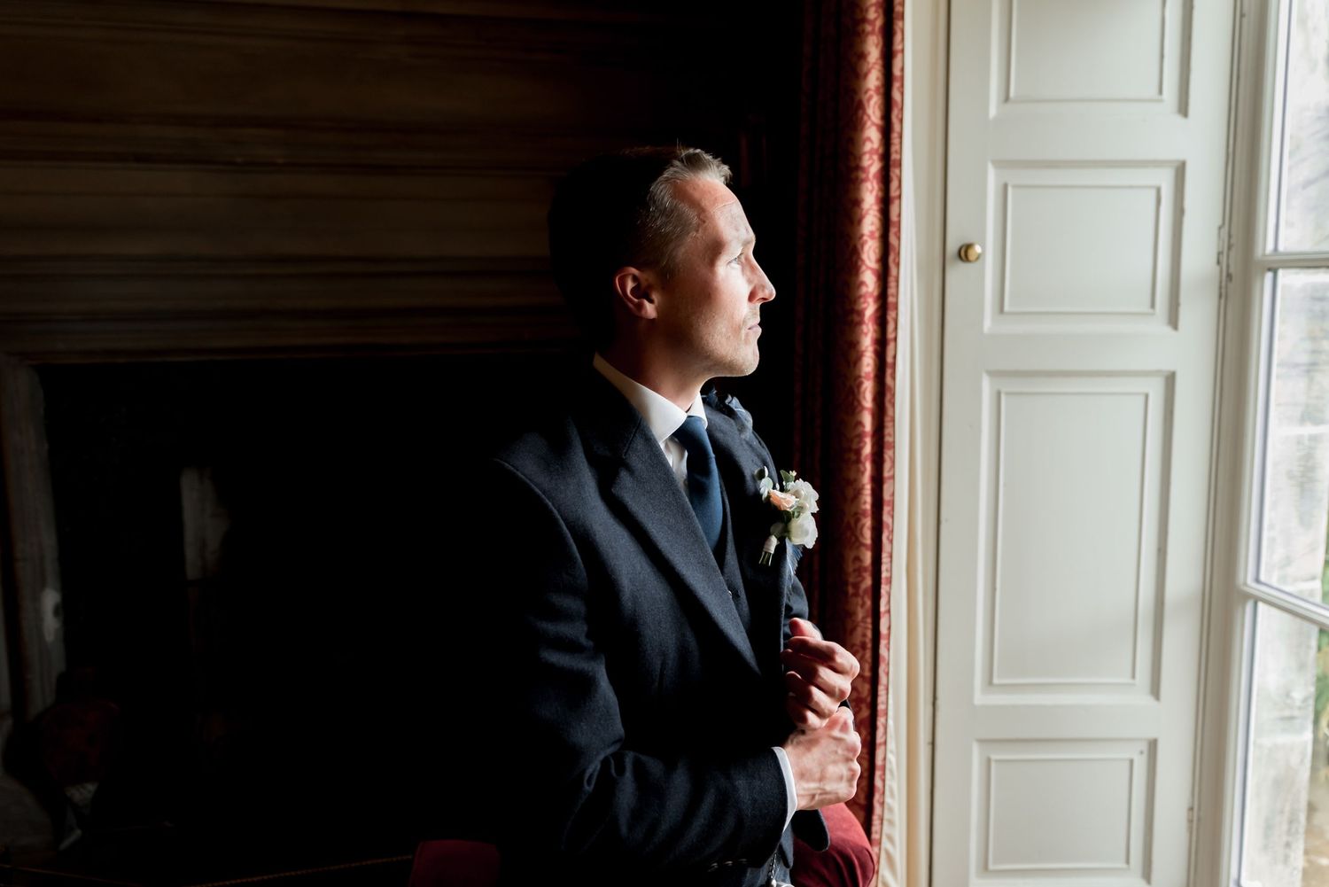 A groom in a dark suit stands by a window in moody lighting, looking contemplatively outside with a boutonniere on his lapel.