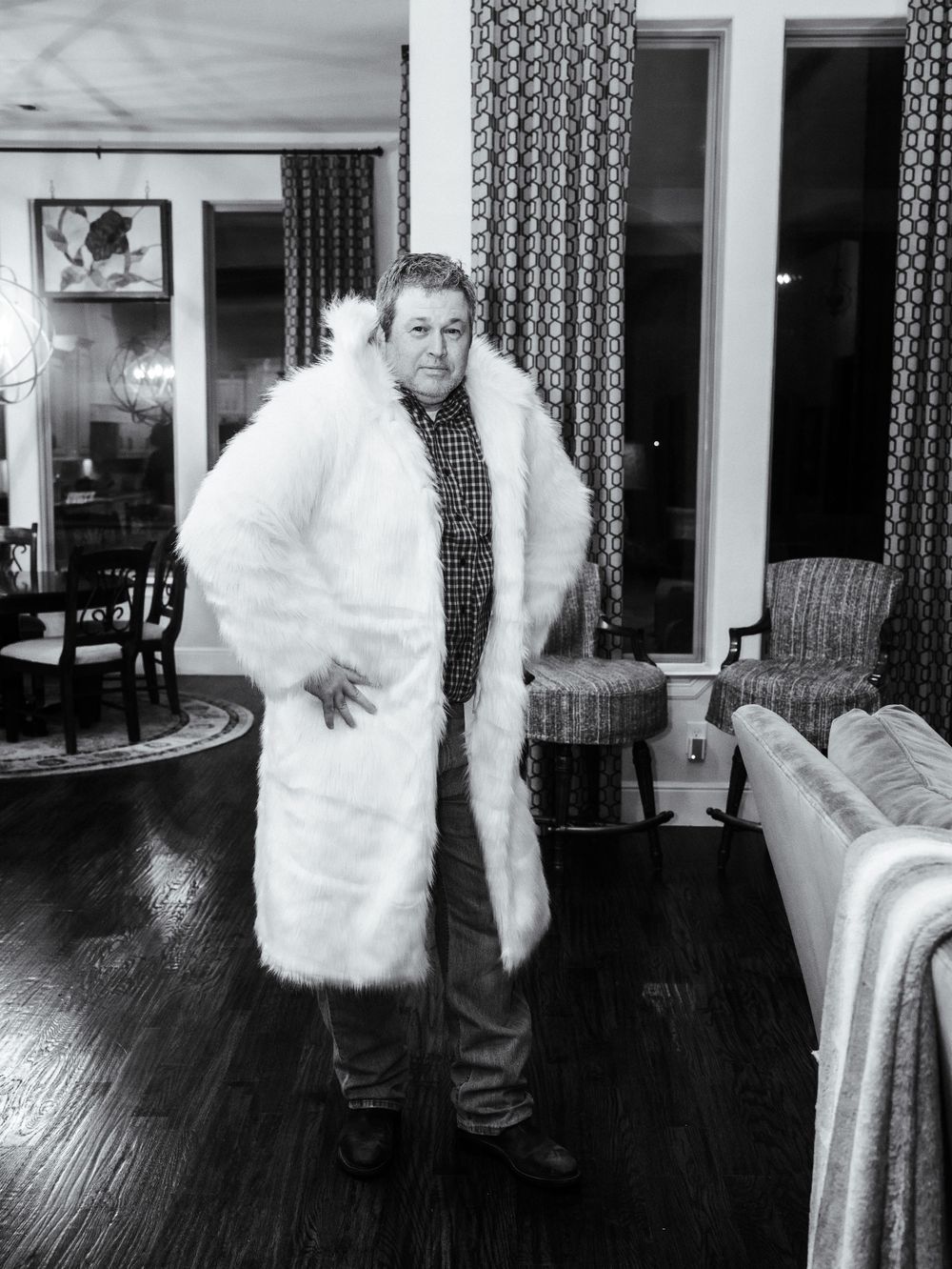A person wearing a long white fur coat and checkered shirt stands in a living room with wood floors and patterned curtains.