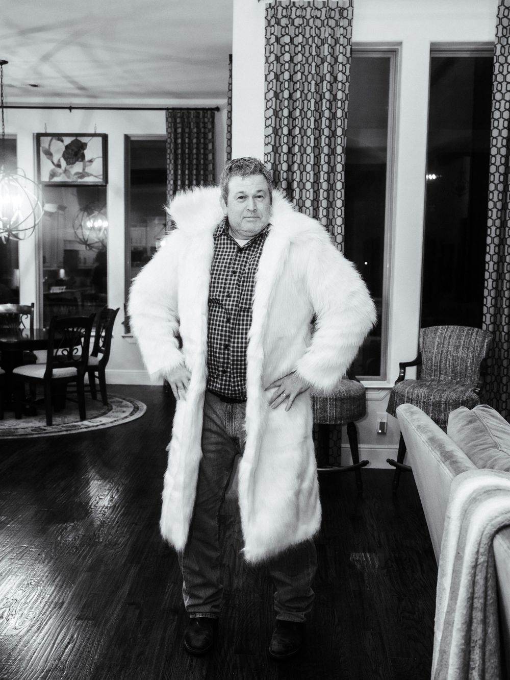 A person poses in a luxurious white fur coat and plaid shirt inside a living room with hardwood flooring.