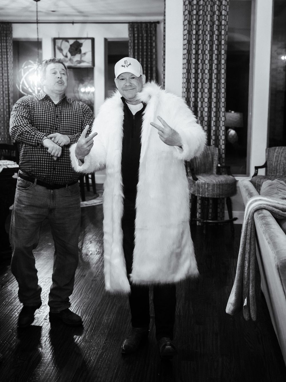 Two people standing indoors, one wearing a casual plaid shirt and the other in a long white fur coat.