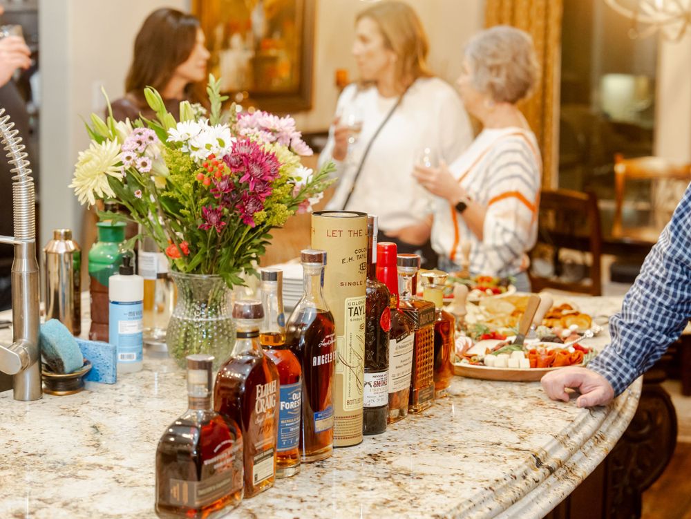 A festive table spread with colorful flower arrangements, various liquor bottles, and plates of food at a party or gathering.