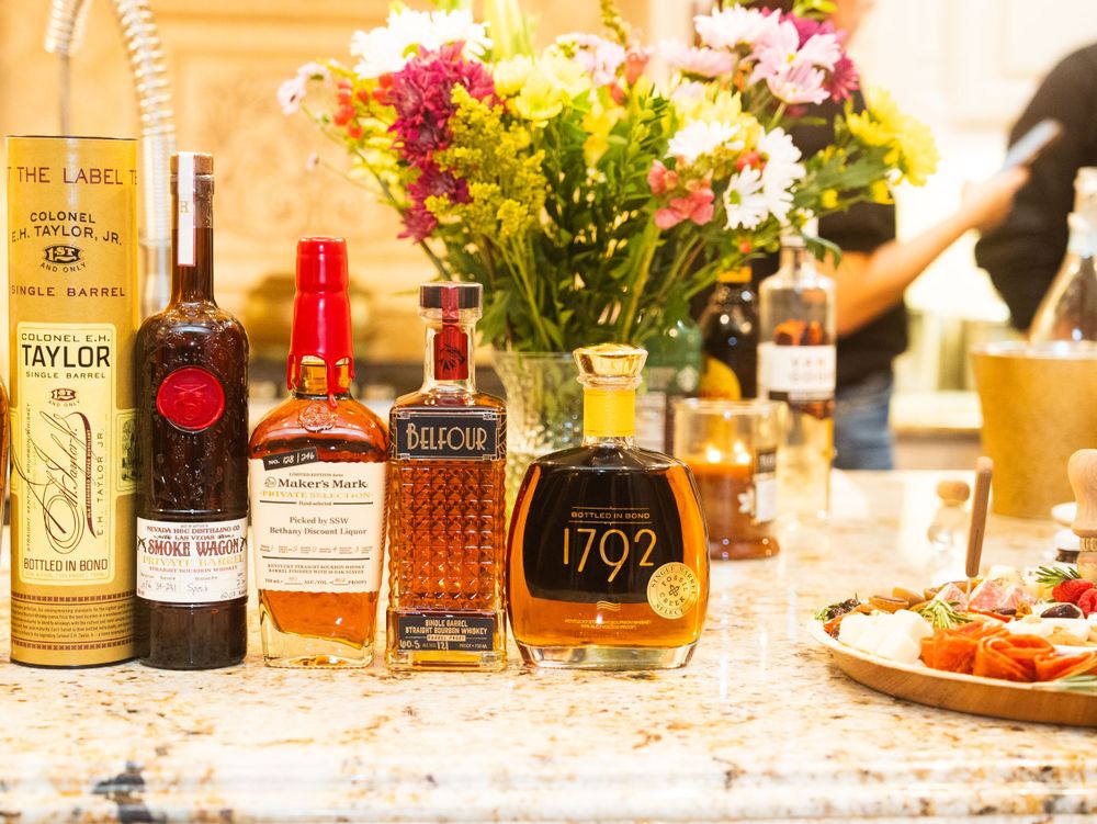 A collection of premium bourbon and whiskey bottles displayed on a counter with colorful flowers in the background.