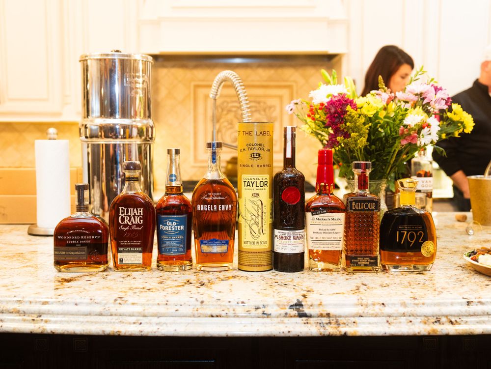 Various bottles of bourbon and spirits lined up on a marble countertop with a vase of colorful flowers in the background.