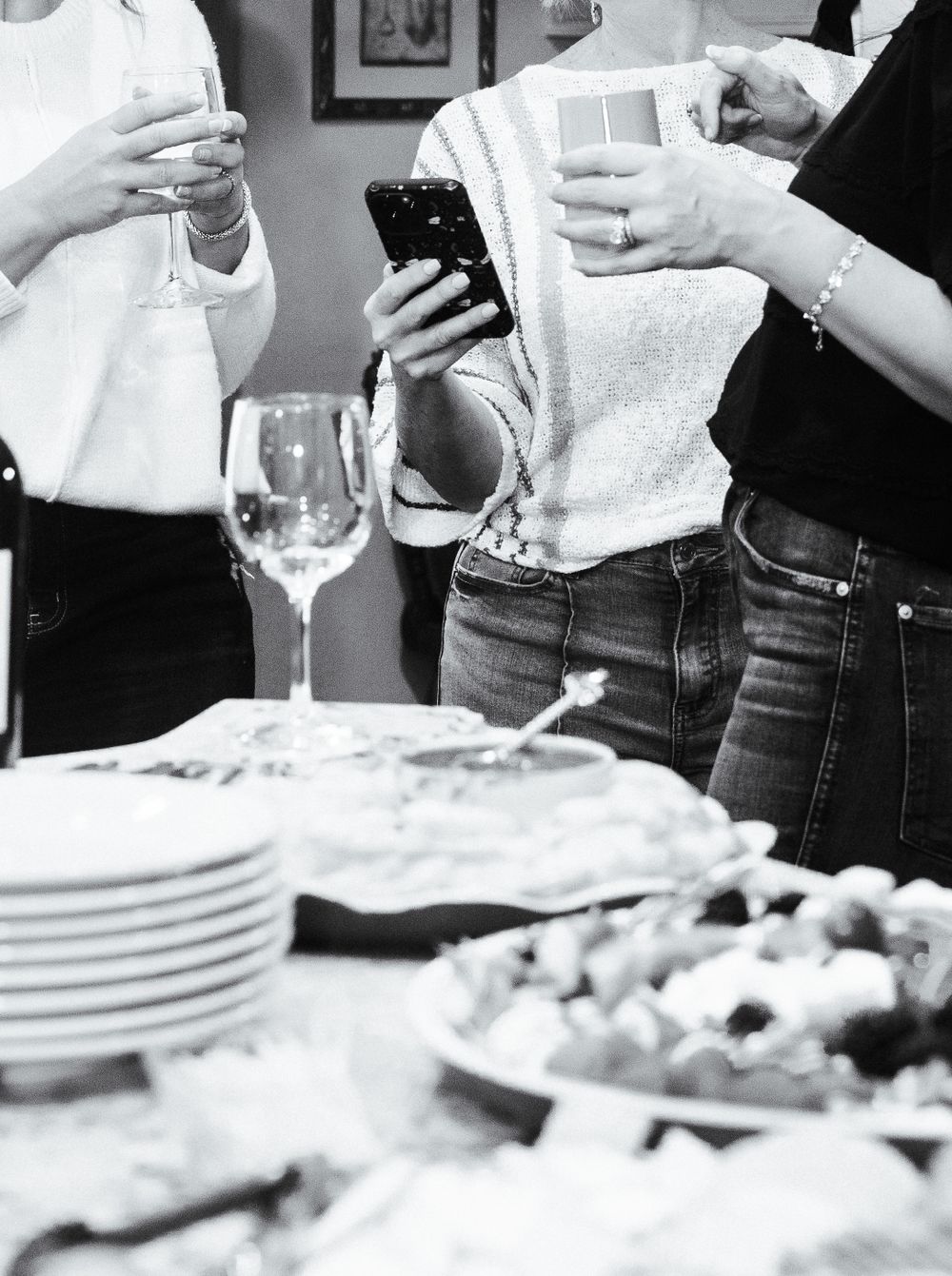 Black and white image of people holding phones and drinks at a party table with appetizers and empty plates.