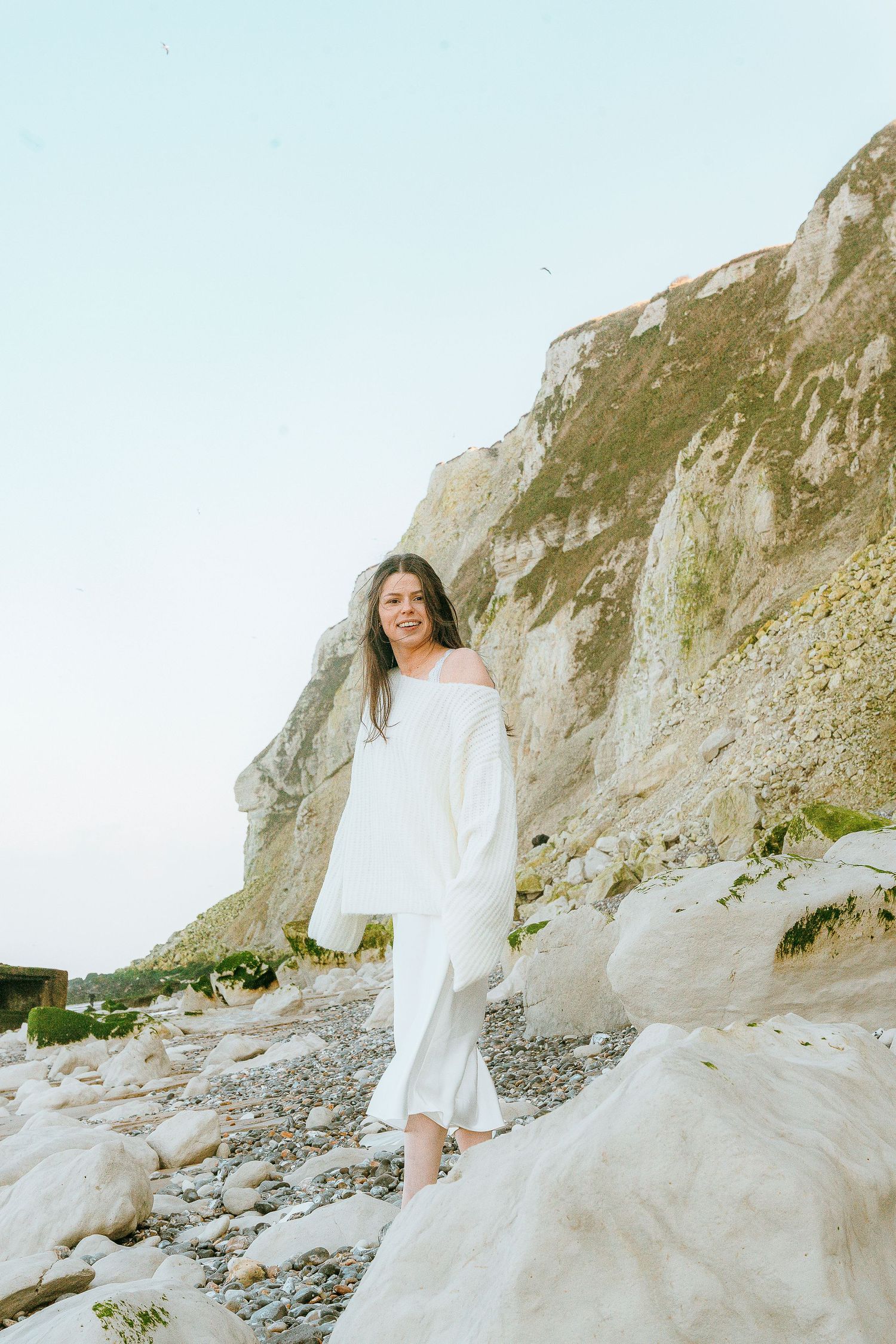 A person in a flowing white dress stands gracefully on rocky coastal cliffs during a bright summer day.