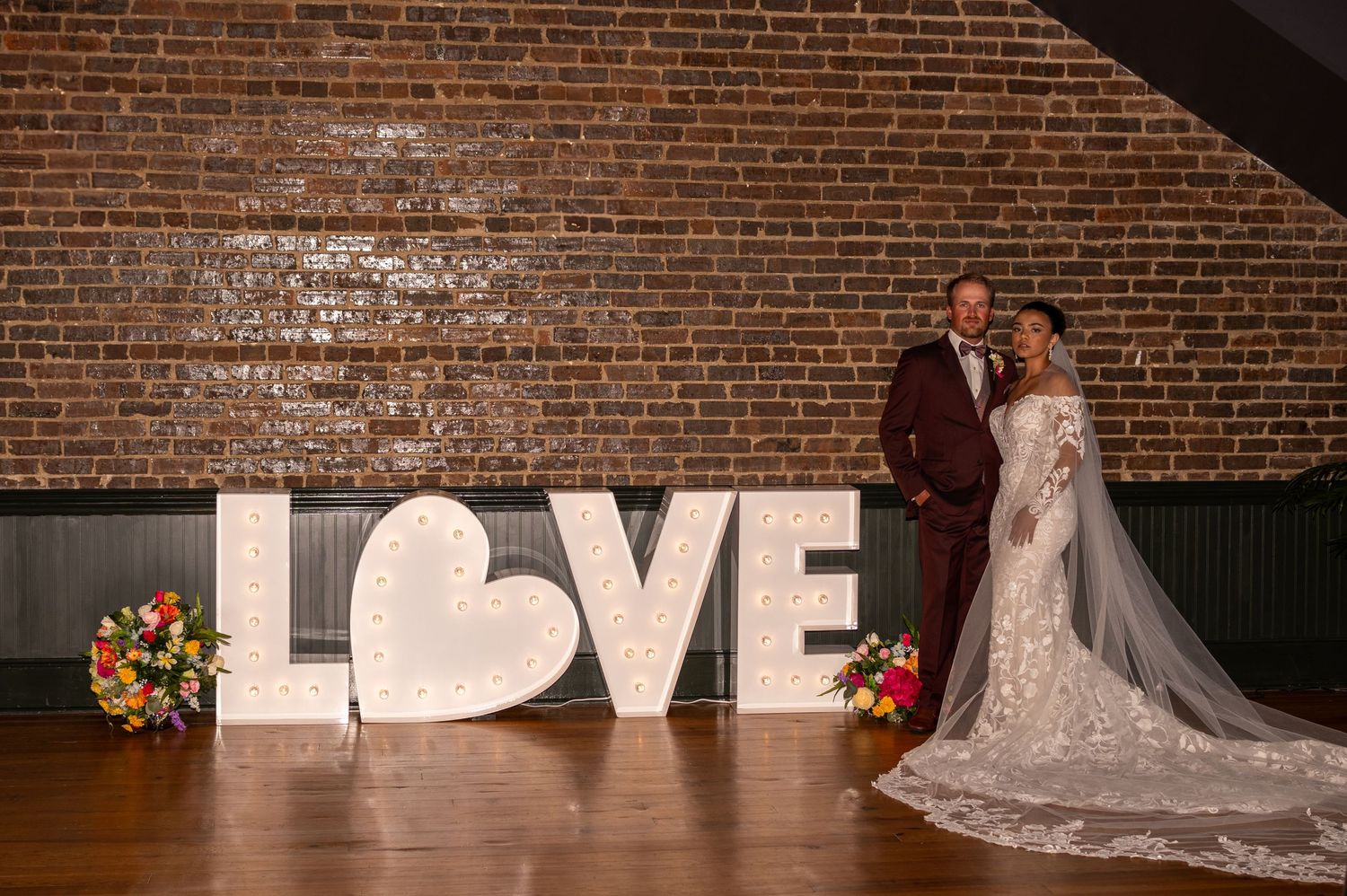 Wedding couple poses in front of large illuminated LOVE letters against rustic brick wall with floral arrangements.