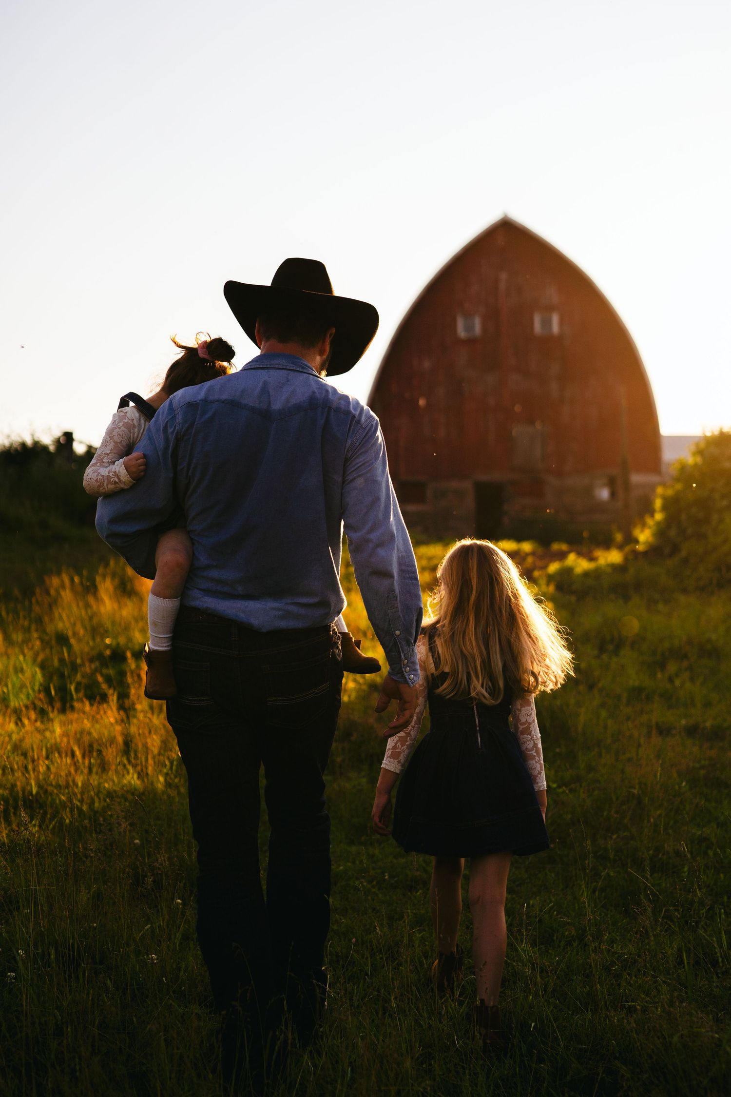 A cowboy with children walks hand in hand toward a rustic barn at sunset in golden rural farmland.