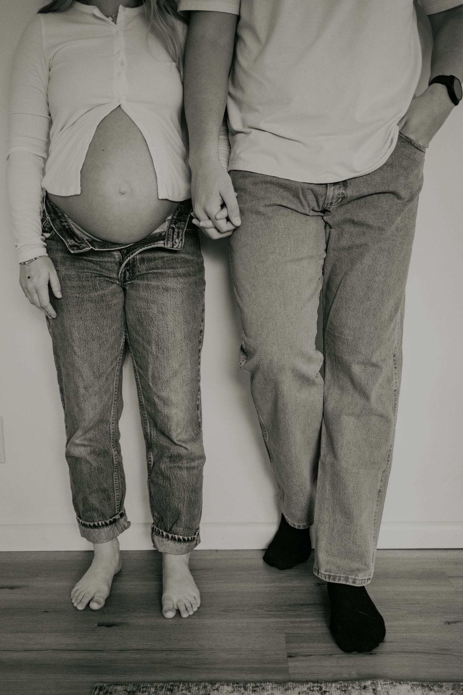 Black and white portrait of a pregnant belly shown in cropped jeans, standing next to partner holding hands.