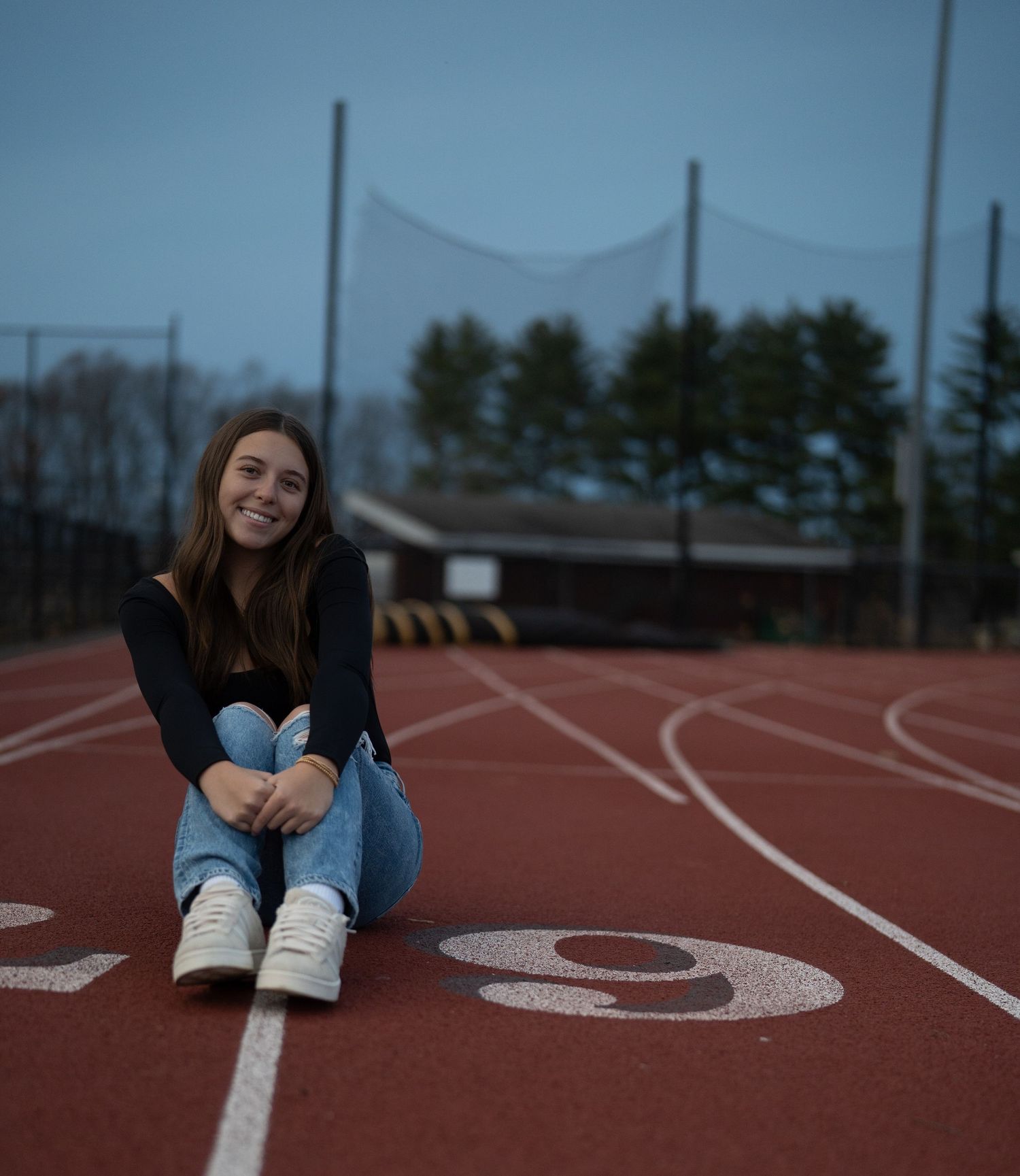 Person sitting on a running track wearing jeans and sneakers at sunset.