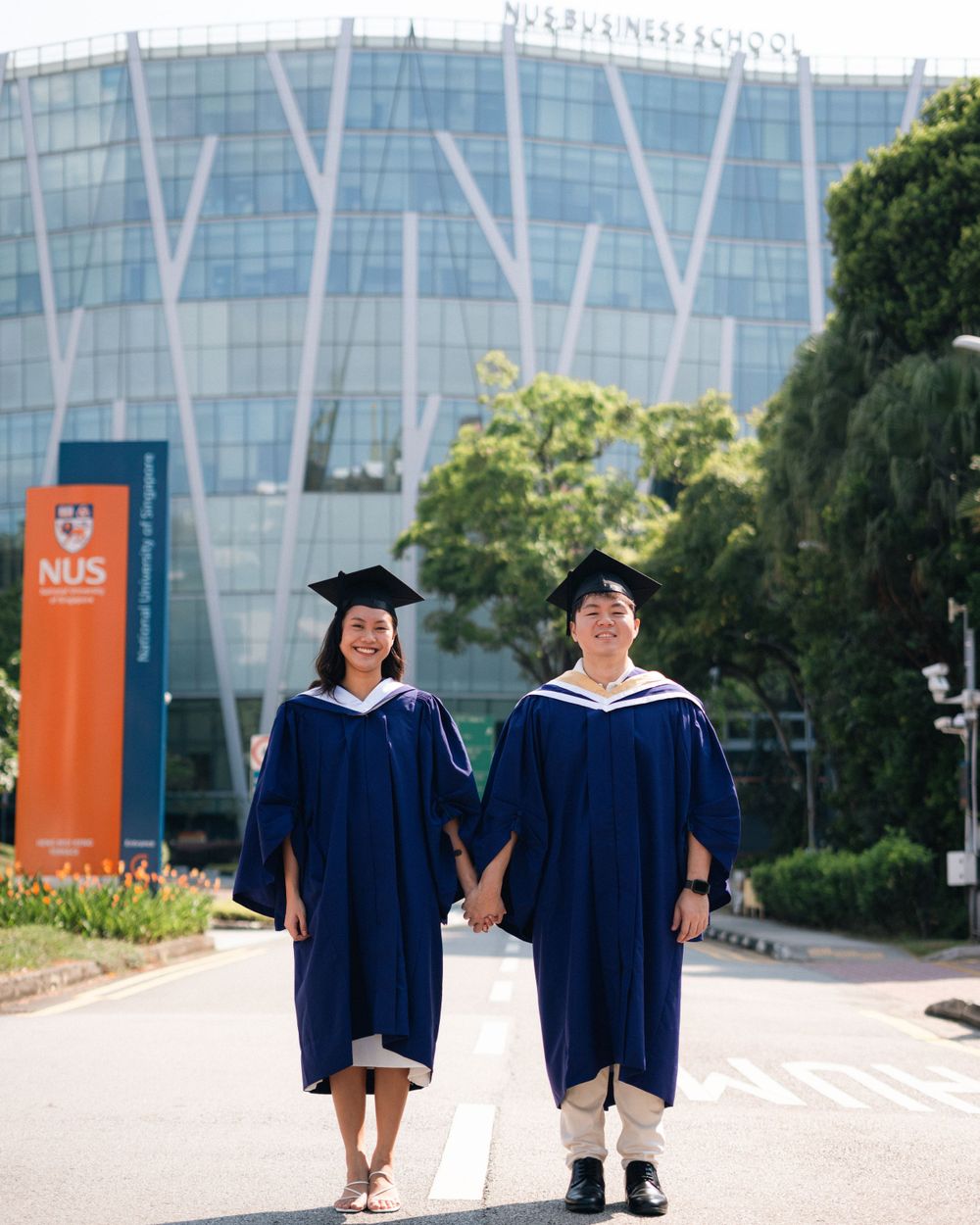 Two graduates in navy robes and mortarboards walking hand in hand on graduation day.