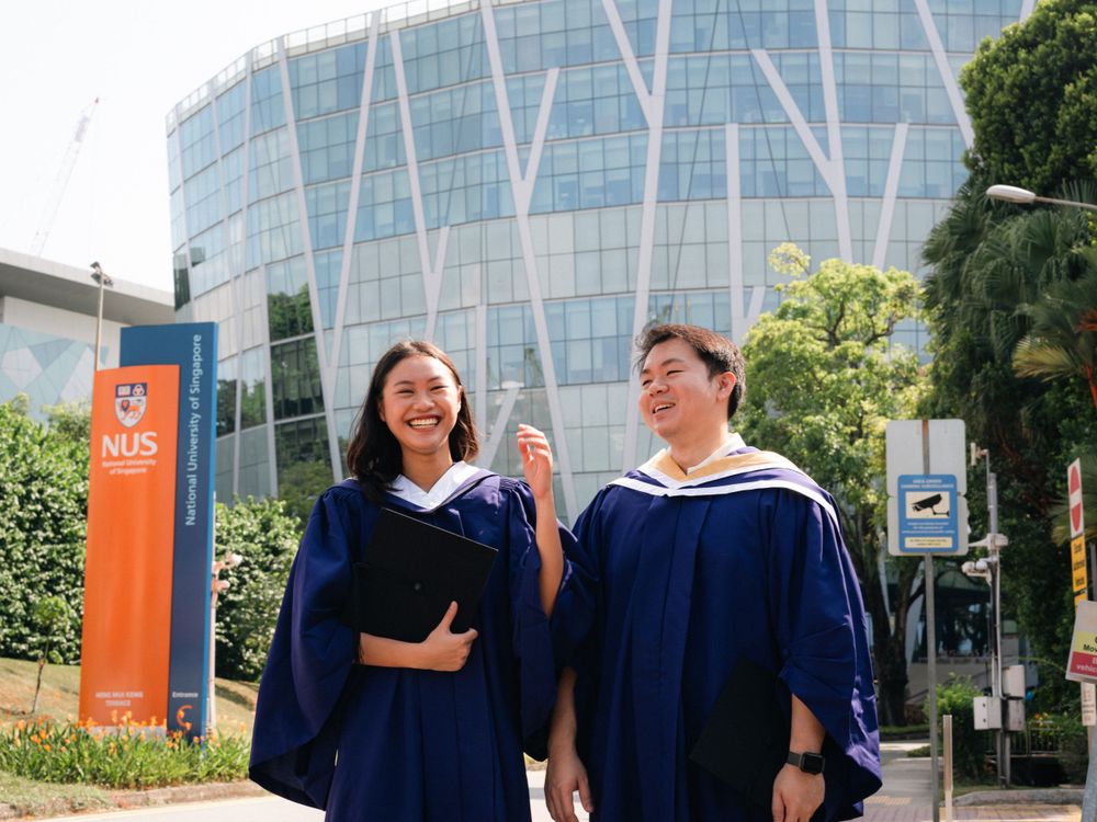 Graduates walk together in blue academic regalia outside a modern glass building with NUS signage.