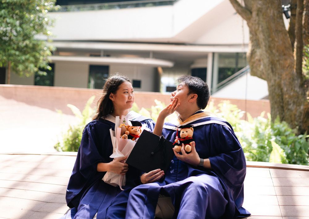 Two graduates in blue robes sitting outdoors sharing snacks and laughing together.