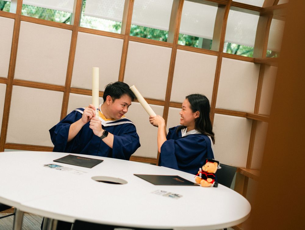 Two graduates playfully posing with their diplomas in an indoor study area.