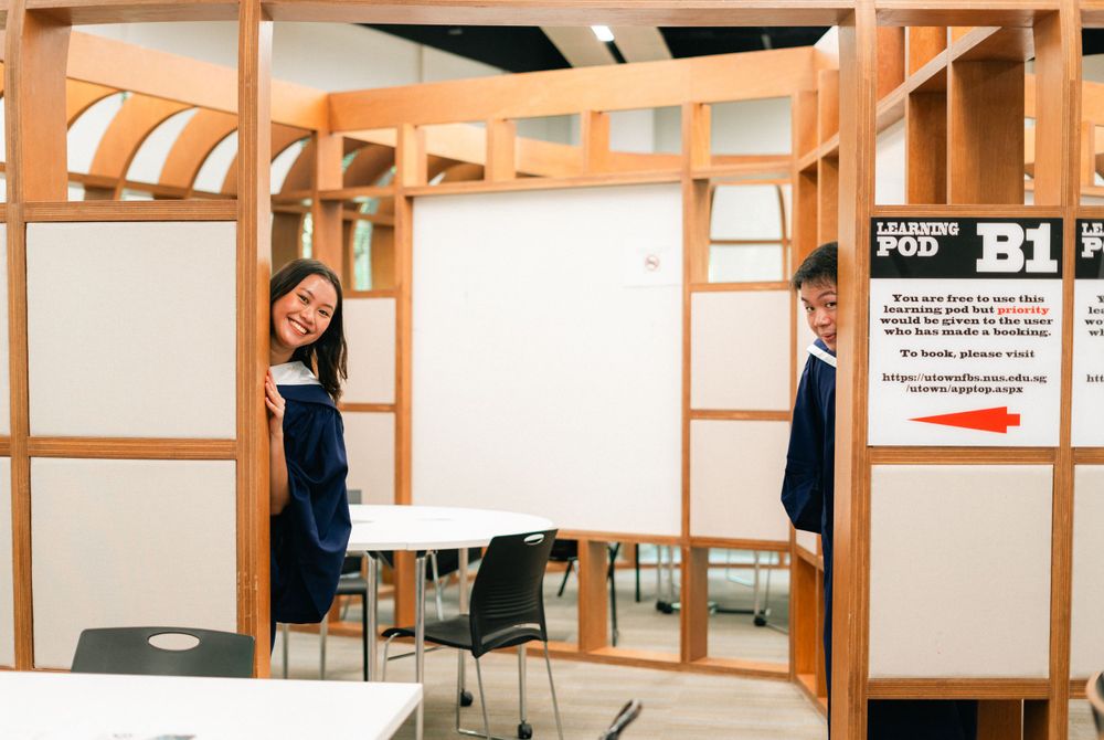 Two graduates playing hide and seek between wooden partitions in a study room.