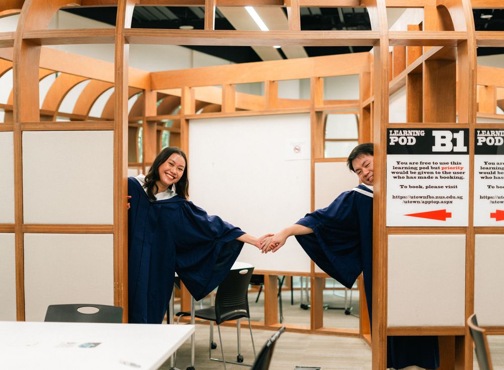 Two graduates holding hands and playing between wooden room dividers.