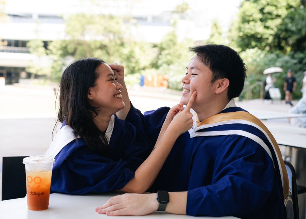 Two graduates in blue robes share a playful moment at an outdoor table.