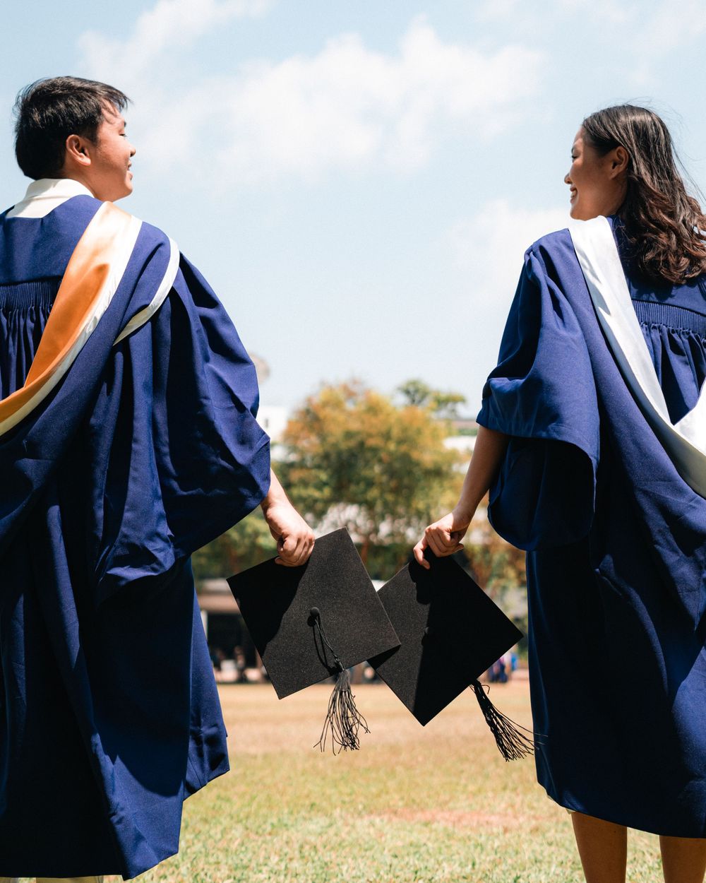 Graduates holding caps while posing back-to-back outdoors.