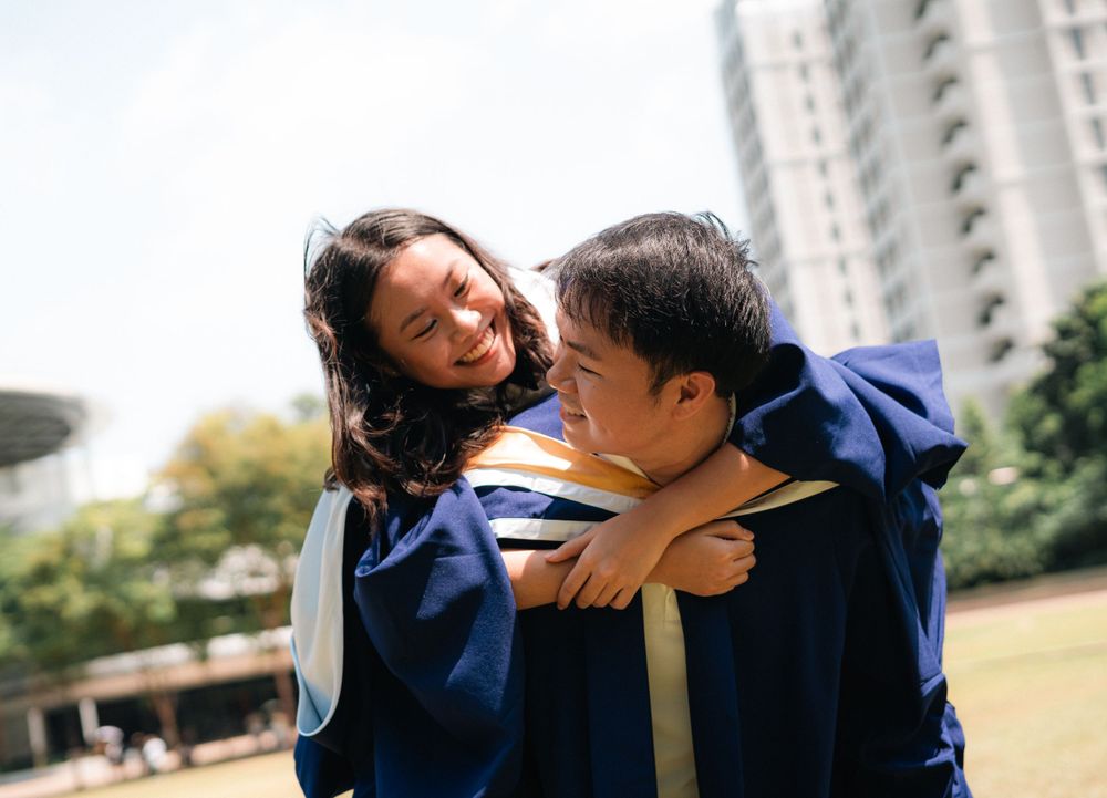 Two graduates in blue academic regalia share a playful moment outdoors on campus.
