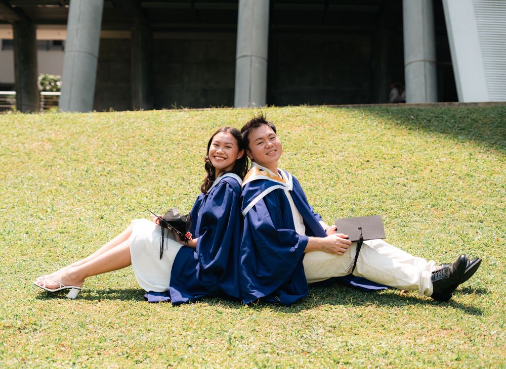 Graduates in blue gowns sitting casually on grass enjoying a lighthearted moment together.