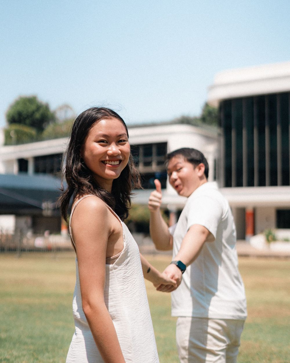 Happy couple in white attire pose playfully on sunny campus grounds.