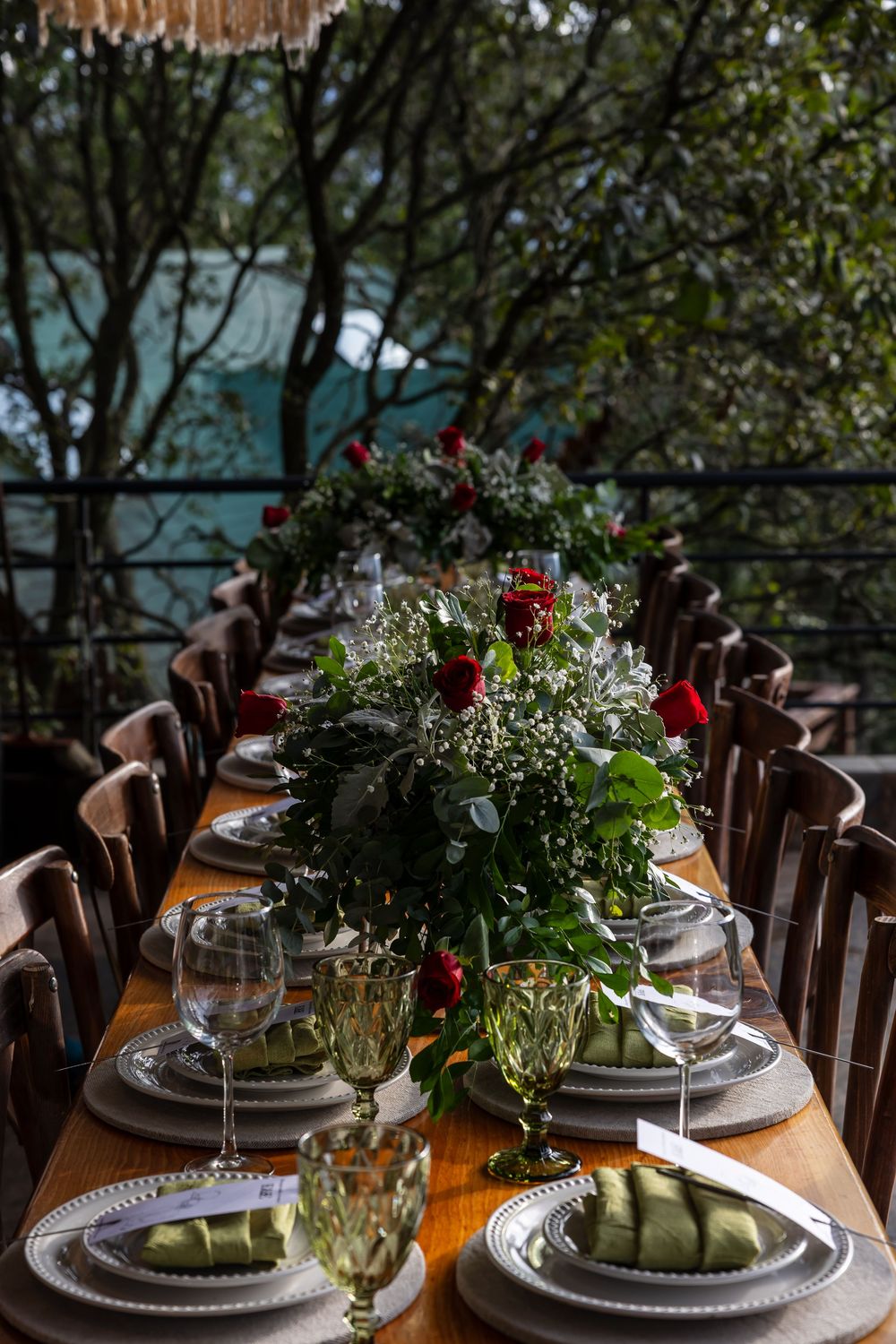 Elegant table setting with white plates, red floral centerpieces and rustic wooden chairs at an outdoor venue.