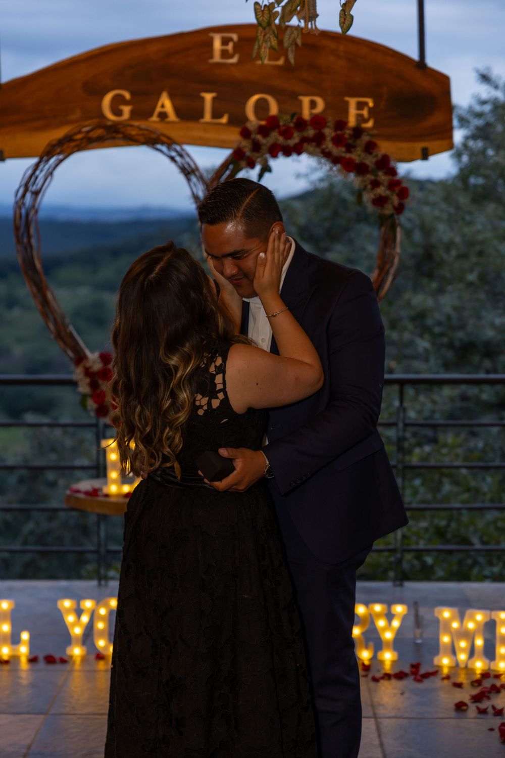 Couple sharing an intimate moment under illuminated wooden signage at twilight.