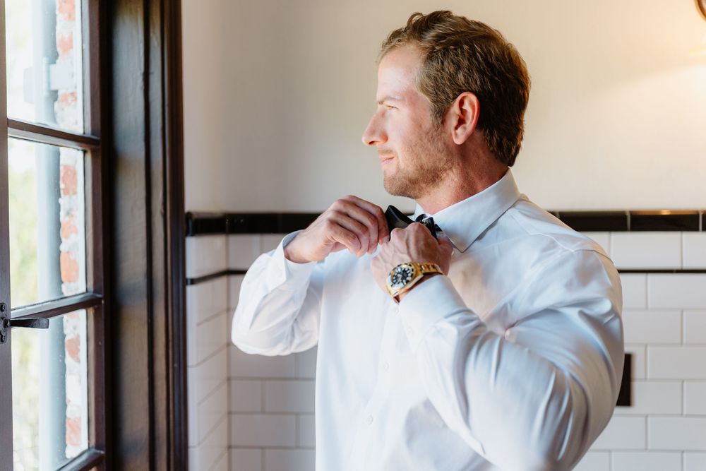 A man in a bow tie and formal attire stands thoughtfully near a window in an elegant interior setting.