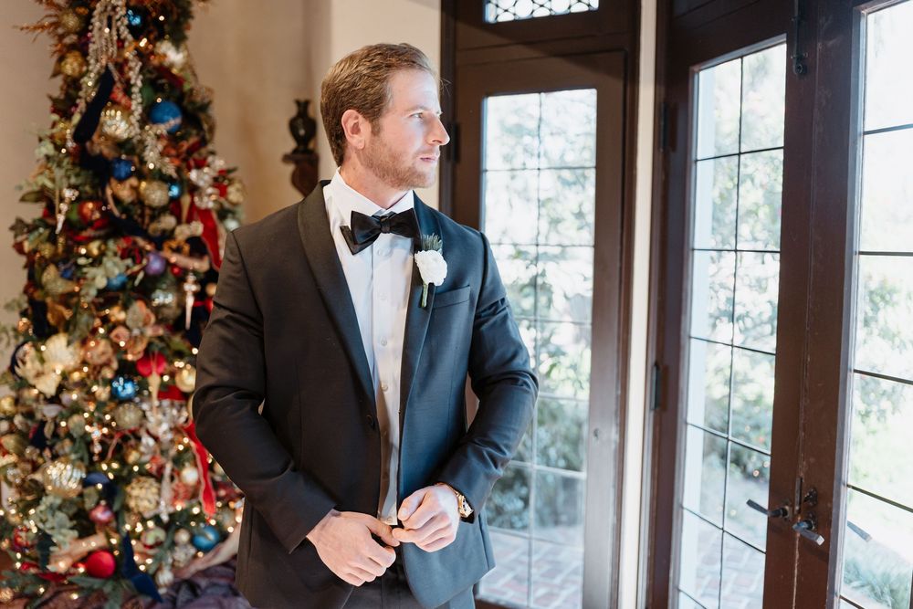 A groom in a black tuxedo and bowtie stands by a decorated Christmas tree near glass doors.