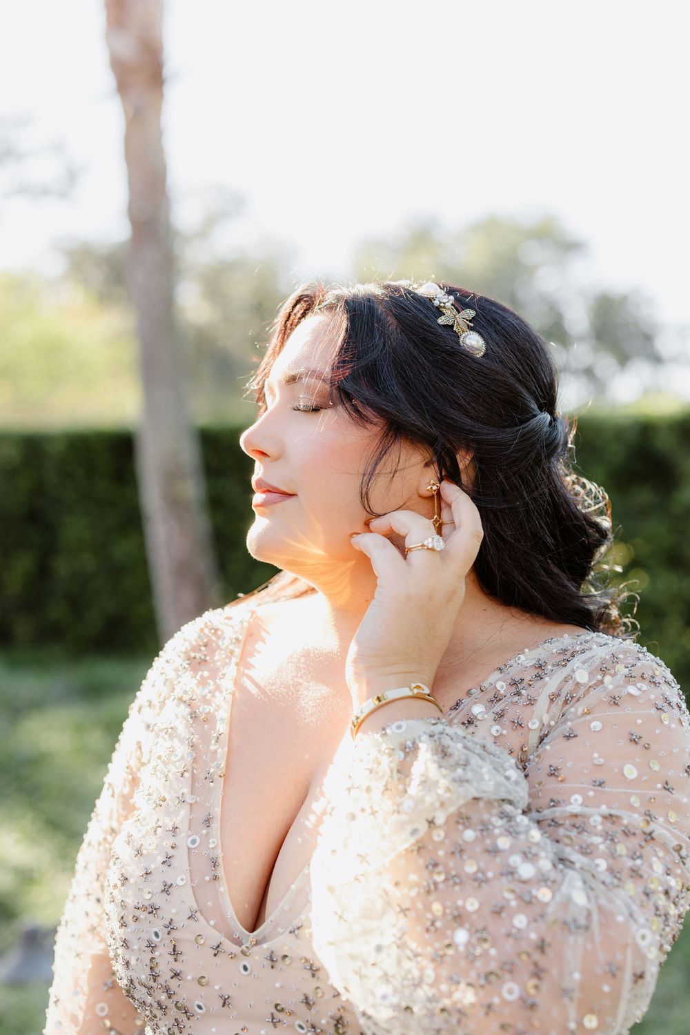 A person in a sheer beaded dress poses outdoors in golden sunlight with eyes closed and head tilted back.