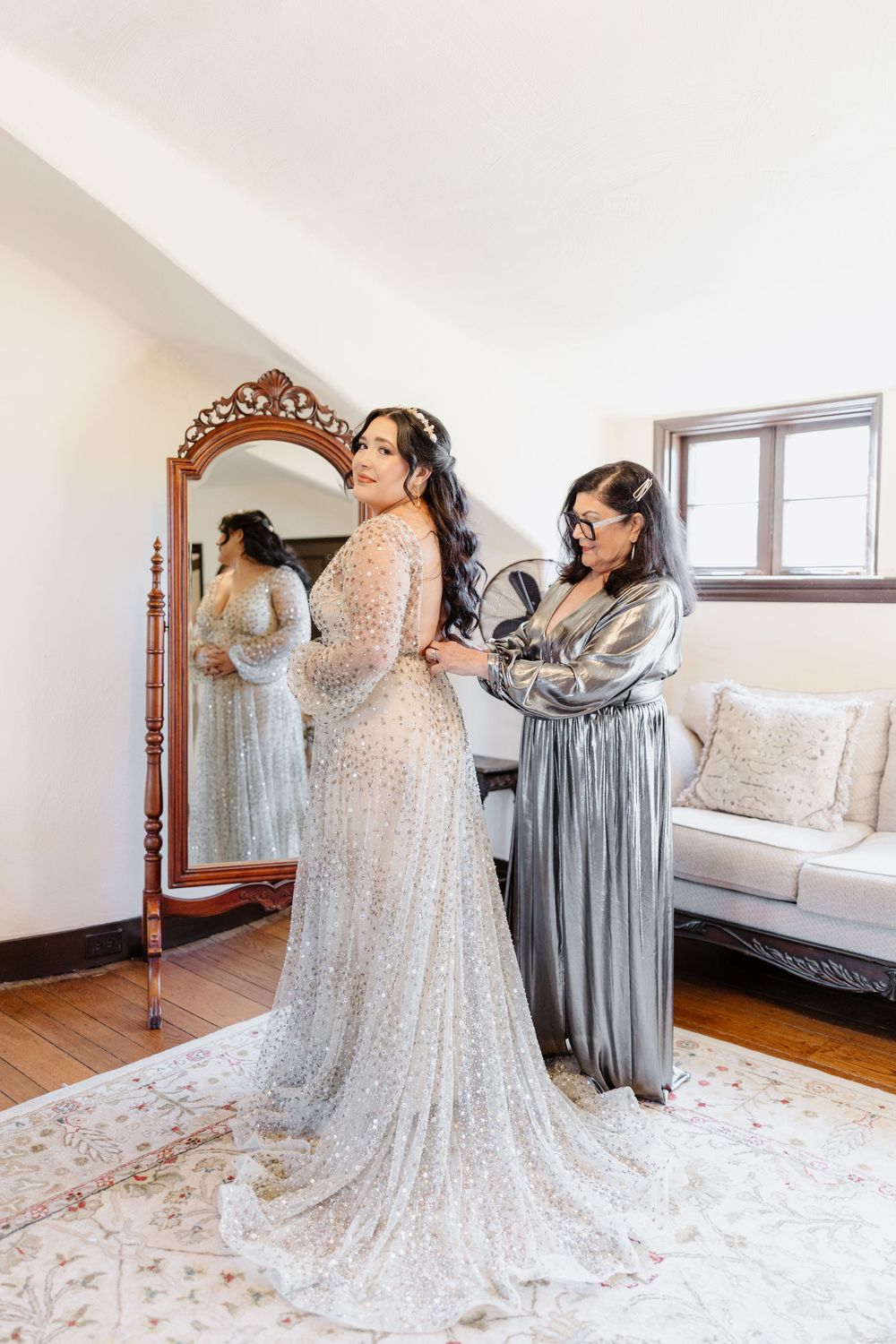 People helping with final wedding dress preparations in a bright sunlit room with hardwood floors.