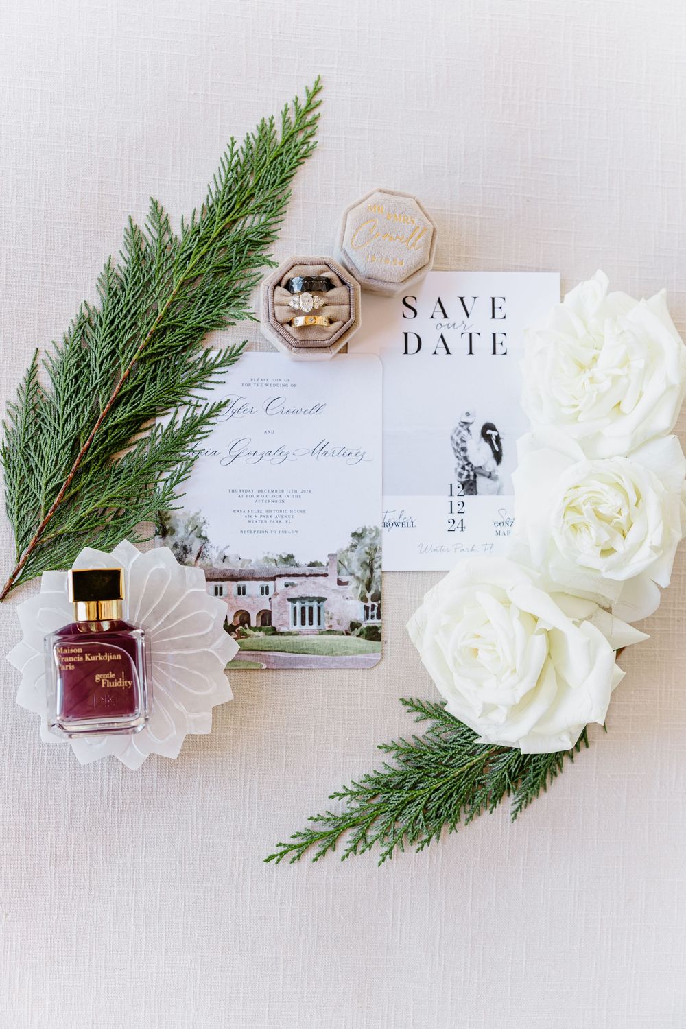 Wedding invitation suite with white roses, evergreen sprigs, and venue illustration laid out in flat lay composition.