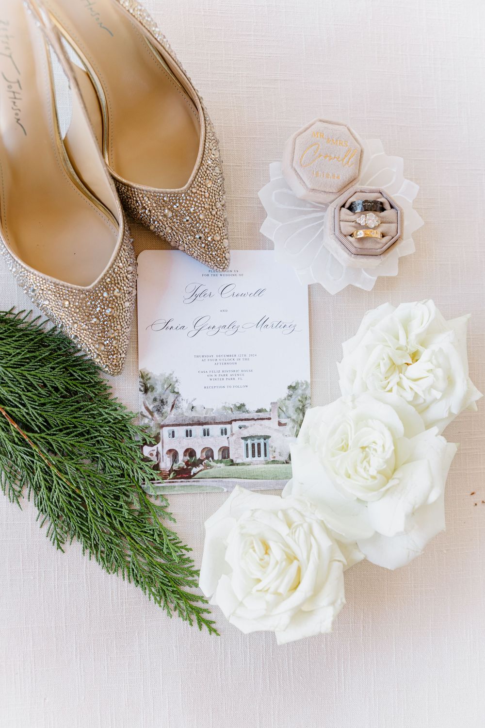 Elegant wedding flatlay with sparkly heels, white roses, evergreen sprigs and invitation on light background.